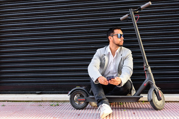 Fototapeta na wymiar Latin adult man with sunglasses, well dressed and electric scooter talking on his mobile phone sitting on the street with a black blind in the background