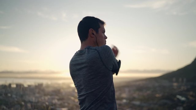 Young fitness man runner wearing earphones stretching hands and back before running on mountain top at sunrise