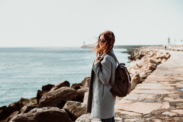 .Pretty young woman with grey coat enjoying a sunny and windy morning around the coast in Porto, Portugal. Walking relaxed, happy and carefree. Lifestyle