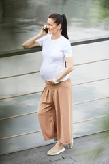 Young stylish pregnant woman standing on the bridge looking at beautiful view of the lake and talking on mobile phone