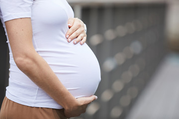 Close-up of young woman in white T-shirt embracing her pregnant belly while standing on the bridge outdoors