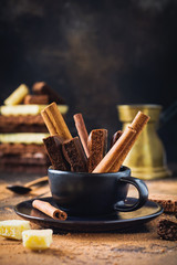 Pieces of porous chocolate and cinnamon sticks in black coffee cup on dark old background. Selective focus.