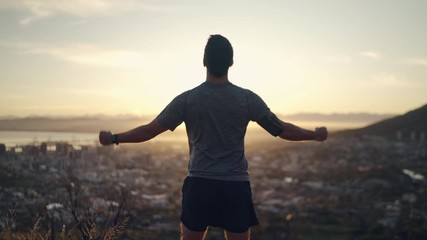Rear view of a sporty healthy male athlete stretching hands with clenched fists celebrating his success in front of rising sun on top of mountain - motivated man celebrating achieving his goals