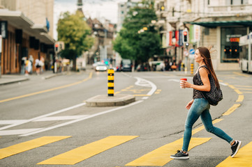 Outdoor portrait of beautiful young woman crossing road in the city, wearing backpack, holding cup...