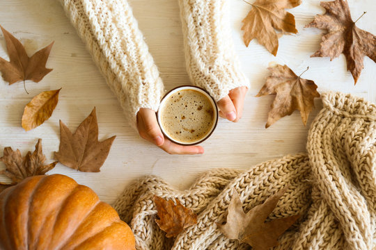 Top view composition with young woman's hands in white sweater, vintage styled cup of coffee and autumn themed decoration, fallen leaves on textured background. Flat lay, copy space.