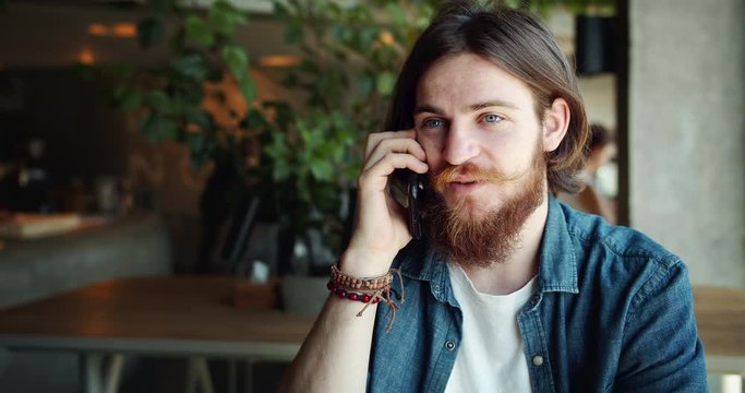 Attractive bearded male hippie talking on smartphone in cafe, communicating with friends, mobile connection