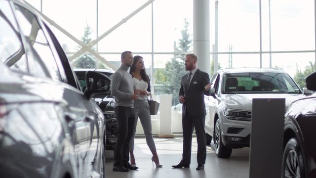 Caucasian family couple listening to salesman and discussing new cars in auto showroom
