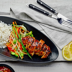Asian- style chicken with savory and sweet teriyaki sauce, rice, sesame, salad, edamame and sweet chili sauce on a wooden table. Top view, directly above.
