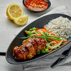 Asian- style chicken with savory and sweet teriyaki sauce, rice, sesame, salad, edamame and sweet chili sauce on a wooden table.