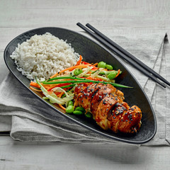 Delicious Asian teriyaki chicken, salad made of cucumber, carrot, ginger, pepper and cabbage, edamame, sesame and rice in a black pottery plate.