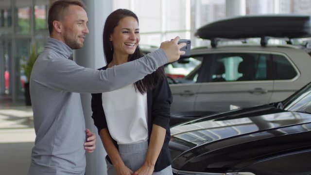 Man taking photo of new car with smartphone and discussing it with happy wife while buying new car in auto dealership showroom