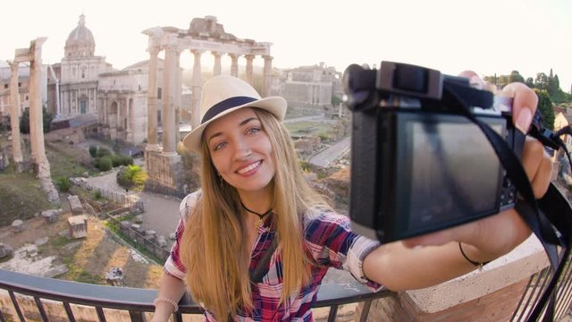 Happy young blonde woman with long hair and blue eyes taking selfies with vintage camera at Roman Forum at sunrise. Rome, Italy