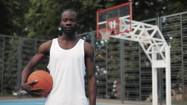 Portrait of Young Muscly Serious Afro - American Guy in White Singlet Holding Ball Standing at Street Basketbal Court and Looking to Camera. Healthy Lifestyle and Sport Concept.