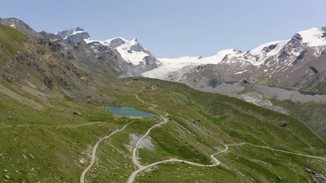 Aerial over mountain roads and path, overflying the Stellisee lake, then climbing towards Findeln glacier and its wide moraines