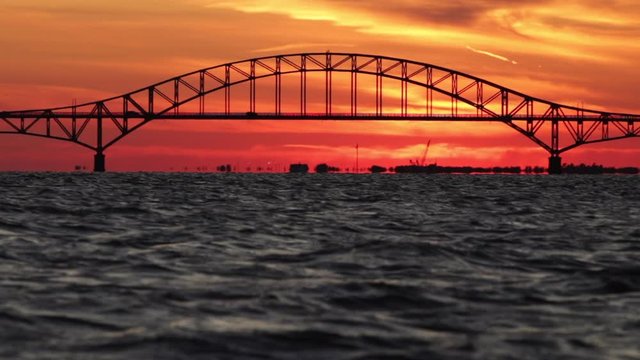 Silhouettes of cars driving over a steel tied arch span bridge at sunset. Vibrant orange sky. 