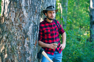 Naklejka na ściany i meble Lumberjack with axe on forest background. Man doing mans job. A handsome young man with a beard carries a tree. Stylish young man posing like lumberjack.