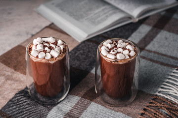 Two glasses of hot cocoa with marshmallows on a brown wool blanket next to an open book