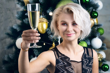 Young blonde woman raises a glass to the New Year celebration sitting in front of a decorated Christmas tree