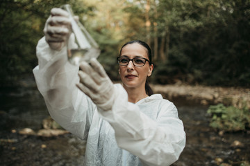 Female scientist biologist and researcher in protective suit taking water samples from polluted river.