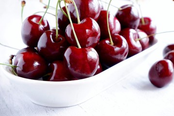 Fresh juicy sweet dark red sweet cherry in a white ceramic bowl on a light wooden background. The concept of a healthy breakfast, a snack. Closeup with selective focus.