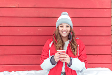 Happy woman smiles, stands background red fence shop sashes, girl winter street weekend city. Warm jacket hat, hand in cup with hot coffee tea, having fun, resting, laughing, free space for text.