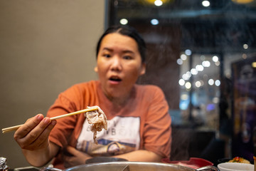 Happy chubby Asian woman holding bamboo chopsticks with a piece of boiled sliced beef enjoying eating in shabu restaurant