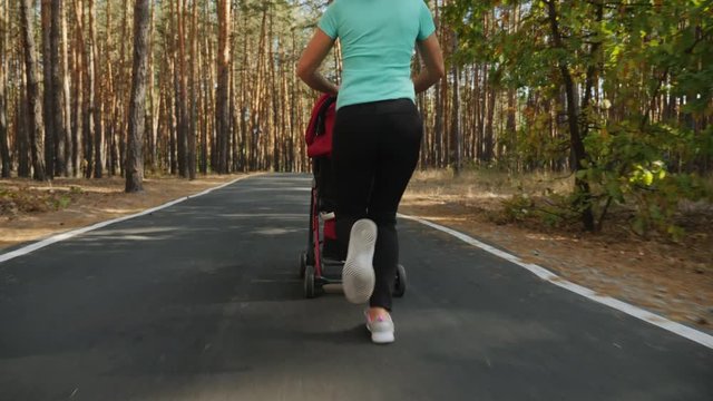Back view: Active woman running with a baby stroller on an asphalt road through a pine forest. Slow-motion 4k video. The camera follows, slowly rises from the bottom up