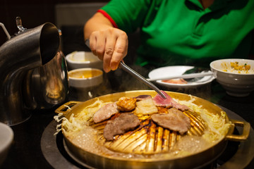 Hand holding tongs with pieces of meat grilling on BBQ brass pan in japanese restaurant