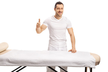 Professional masseur standing behind a massage bed and showing thumbs up - obrazy, fototapety, plakaty