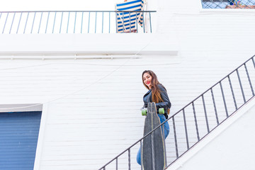 smiling young woman with long brown hair, going down stairs with a longboard in her hand, with jeans pants and leather jacket on a white brick background