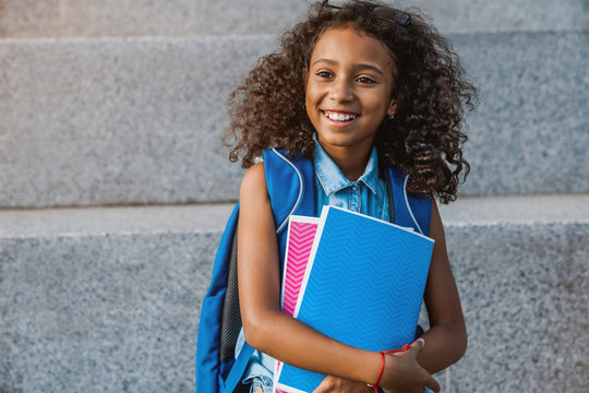 Portrait Of Happy African School Girl With Backpack Holding Notebooks Outside The Primary School