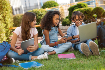 Happy little school children with digital gadgets and backpacks sitting at the grass park outdoors