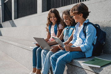 Happy group of multiethnic school kids sitting and using digital devices outdoors