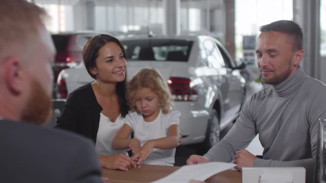 Happy caucasian family buying new car in auto showroom: man giving signed contract to salesman, receiving key and speaking with happy wife and little daughter