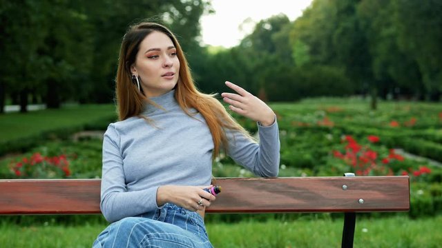 Portrait of redhead of women smokes vape electronic cigarette and blows steam from mouth, rest on bench in summer sunny park