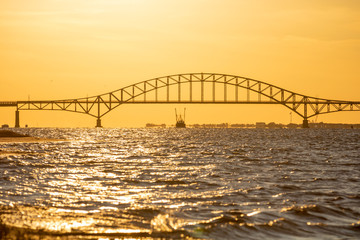 Fishing boat sailing under a steel tied arch span bridge during sunset, with golden light shining over the water. Great South Bay, Long Island New York.