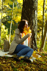 A young brunette girl in jeans and a coat sits in an autumn park on a plaid and holds a maple leaf, looks to the side, near a leather backpack