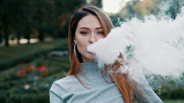 Close-up portrait of redhead of women smokes vape electronic cigarette and blows steam from mouth, rest in summer sunny park