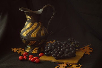 Black and gold vintage jug next to black small grapes on a wooden board. A row of yellow oak leaves and rose hips. Still life.