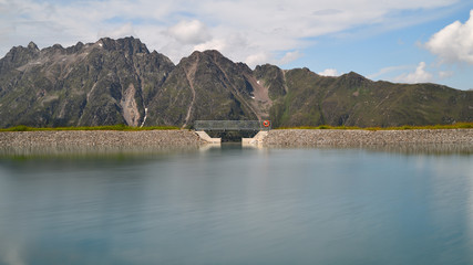 Smooth waters of an alpine water reservoir high up in the Tyrolean mountains in Austria. A modern footbridge with attached red lifebelt crosses the lake. 