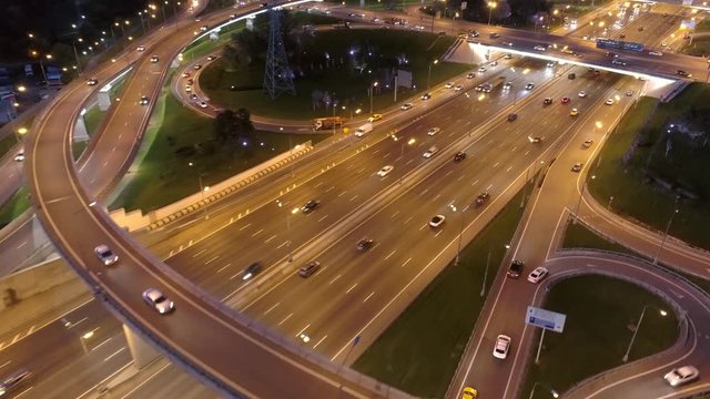 Flight low over night road junction multi-level overpass multi-lane active traffic. Follow cars with headlights on go along an illuminated highway. Transportatian large city Cinematic aerial 4K