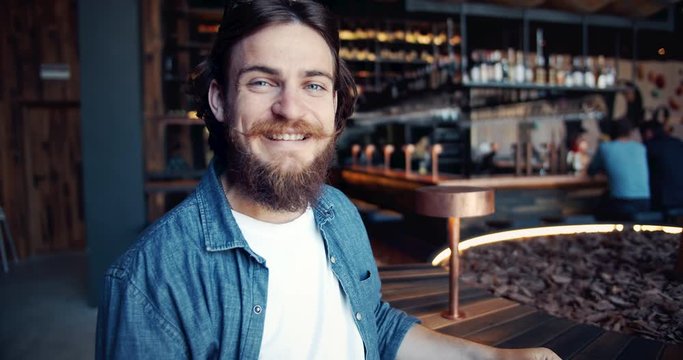 Handsome young male hipster in denim shirt twisting moustache and smiling to camera, trendy man portrait