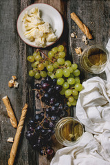 Swiss cheese tete de moine, black and green grapes and two glasses of white wine, served with bread sticks and cloth on old wooden plank background. Flat lay, space