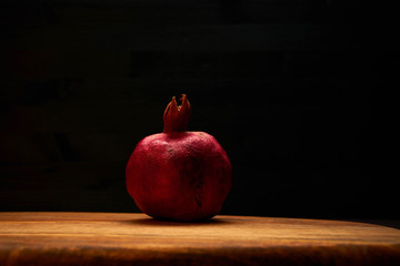Dark red pomegranate on textured medium gold tone wooden chopping board against dark wooden background. Soft warm light from above, front view. Punica granatum