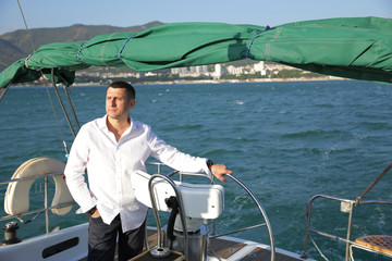 attractive man in white stands at the helm of a yacht at sea. Sailing, tourism, travel and people concept. Happy man at the on steering wheel and navigation.