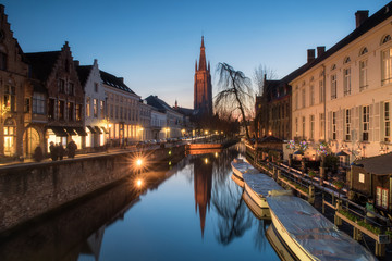 Church of our lady  with traditional building facade reflects on still canal with tourist boats foreground during twilight at Bruges, Belgium