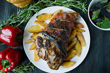 Fried pork knuckle with potatoes served on a white plate. Decorated with fresh Bulgarian pepper, rosemary. Dark wooden background. View from above.