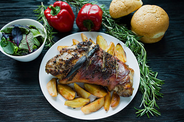 Fried pork knuckle with potatoes served on a white plate. Decorated with fresh Bulgarian pepper, rosemary. Dark wooden background. View from above.
