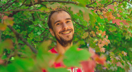 A man in the Park smiles standing behind the leaves. Portrait of a young man. Standing behind bushes and smiles