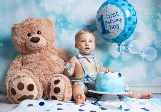 First Birthday: Young Toddler Posing For Messy Cake Smash Photoshoot With Teddy Bear And Balloon In Front Of Blue Decorated Background.